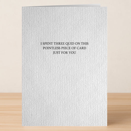 The Pointless Funny Greeting Card by Twisted Gifts features bold black text: "I spent three quid on this pointless piece of card just for you" and is displayed on a wooden surface.