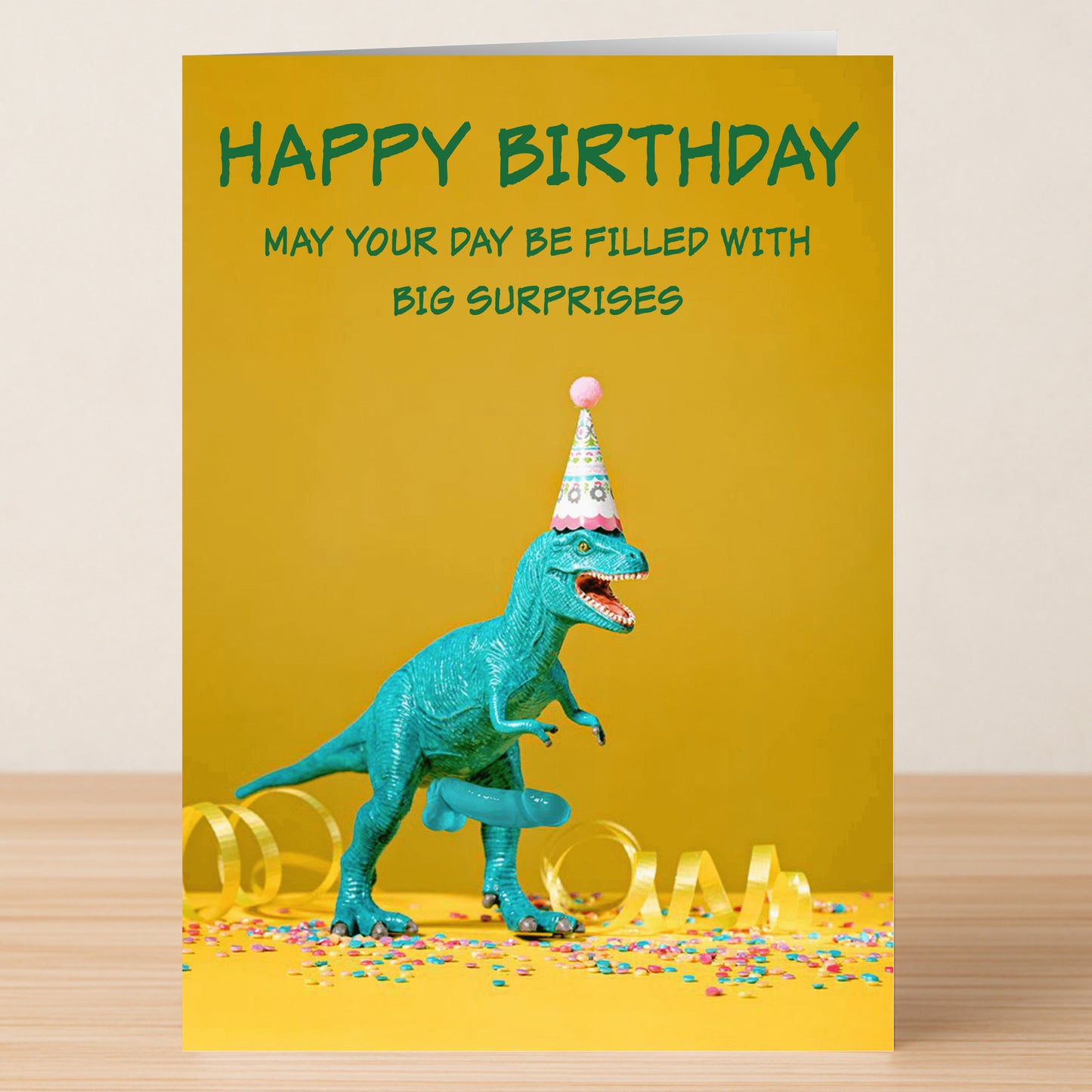 A green toy dinosaur in a party hat stands on yellow, surrounded by confetti, in front of Twisted Gifts’ “Big Suprise Rude Birthday Card.”.