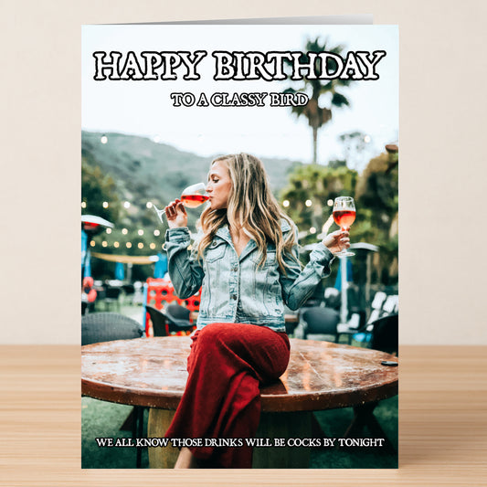 A woman in a denim jacket and red skirt sits outdoors holding two drinks. She has the "Classy Bird Insulting Birthday Card" by Twisted Gifts, featuring witty, light-hearted birthday humor about drinks.