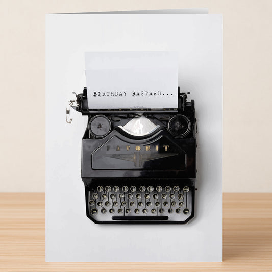 The Bastard Insulting Birthday Card by Twisted Gifts features a black vintage typewriter with "BIRTHDAY BASTARD..." on paper, resting on wood—all against a plain white backdrop, showcasing witty wordplay.