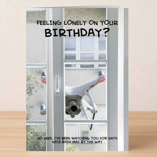 A white drone hovers outside a window; text above reads "Feeling lonely on your birthday?" with a cheeky message below about the drone watching. Perfect for anyone who’d love Twisted Gifts’ Bath Mat Rude Birthday Card.