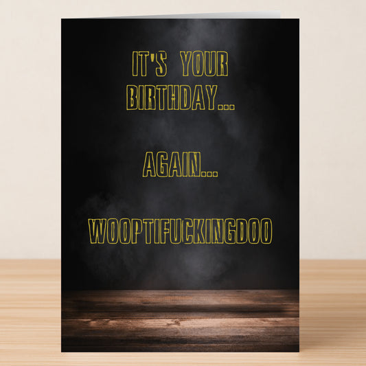 The Woopti Insulting Birthday Card by Twisted Gifts features a black smoky background, wooden surface, and bold yellow-outlined text: "IT'S YOUR BIRTHDAY... AGAIN... WOOPTIFUCKINGDOO"—a funny card that says it all!.