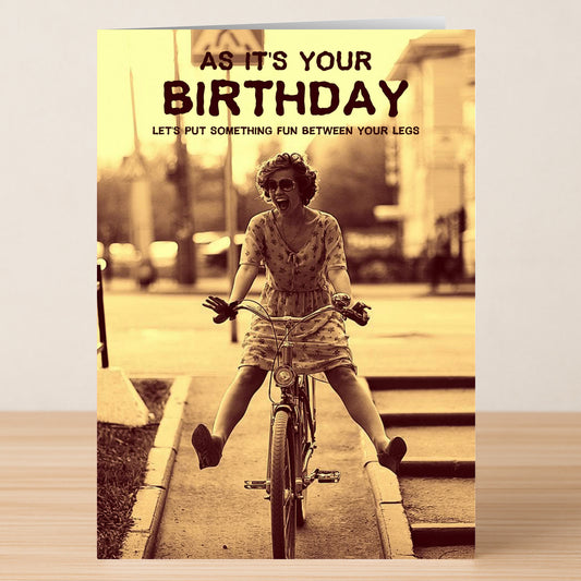 A woman in a dress bikes happily with the text, "As it's your birthday, let's put something fun between your legs." The Fun Rude Birthday Card by Twisted Gifts is a cheeky way to make someone laugh on their special day!.