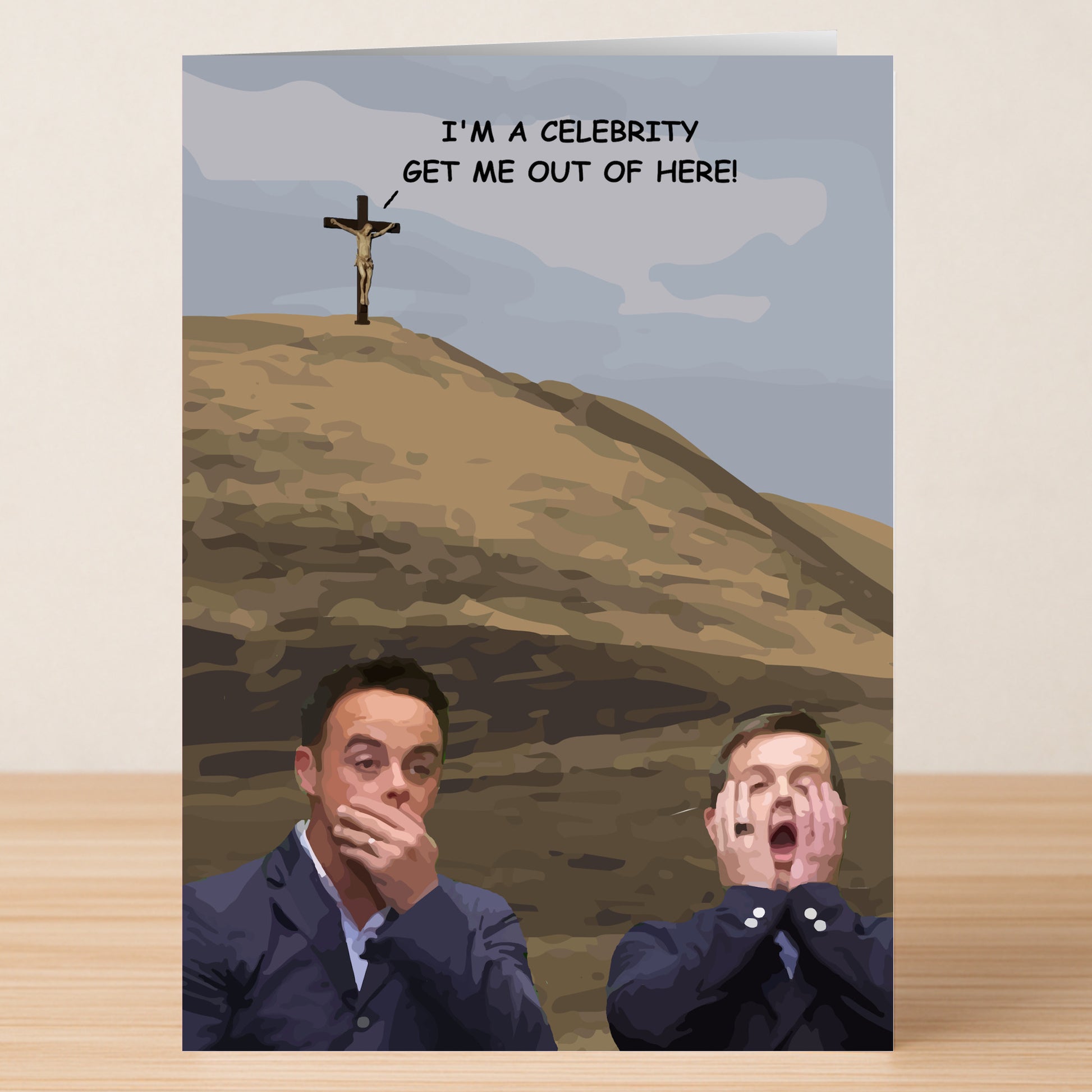 Two men pull shocked faces before a hillside crucifixion scene, with "I'M A CELEBRITY GET ME OUT OF HERE!" above—Twisted Gifts’ I’m A Celebrity Funny Christmas Card delivers an unforgettable and hilarious holiday moment.