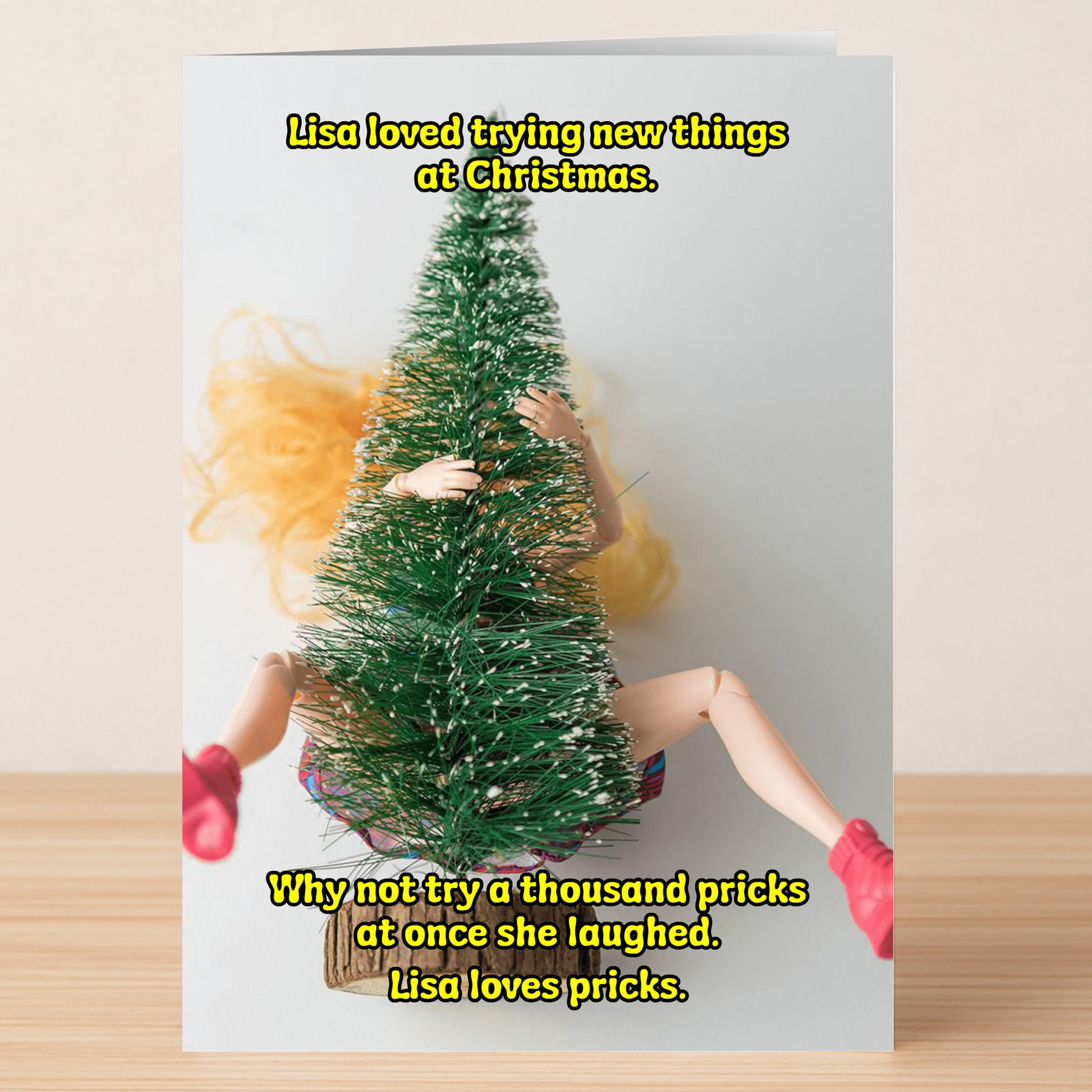 The Loves Pricks Rude Christmas Card by Twisted Gifts features a blonde doll in pink shoes hidden behind a small fake Christmas tree, with funny yellow text about trying new things—great for quirky holiday greetings.