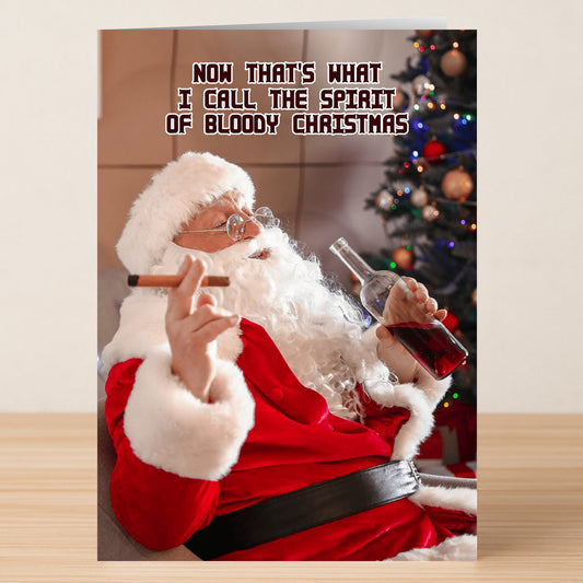 The Spirit Funny Christmas Card by Twisted Gifts features Santa holding a cigar and bottle in front of a Christmas tree with the caption, "Now that's what I call the spirit of bloody Christmas." A humorous holiday card sure to spark festive laughter.