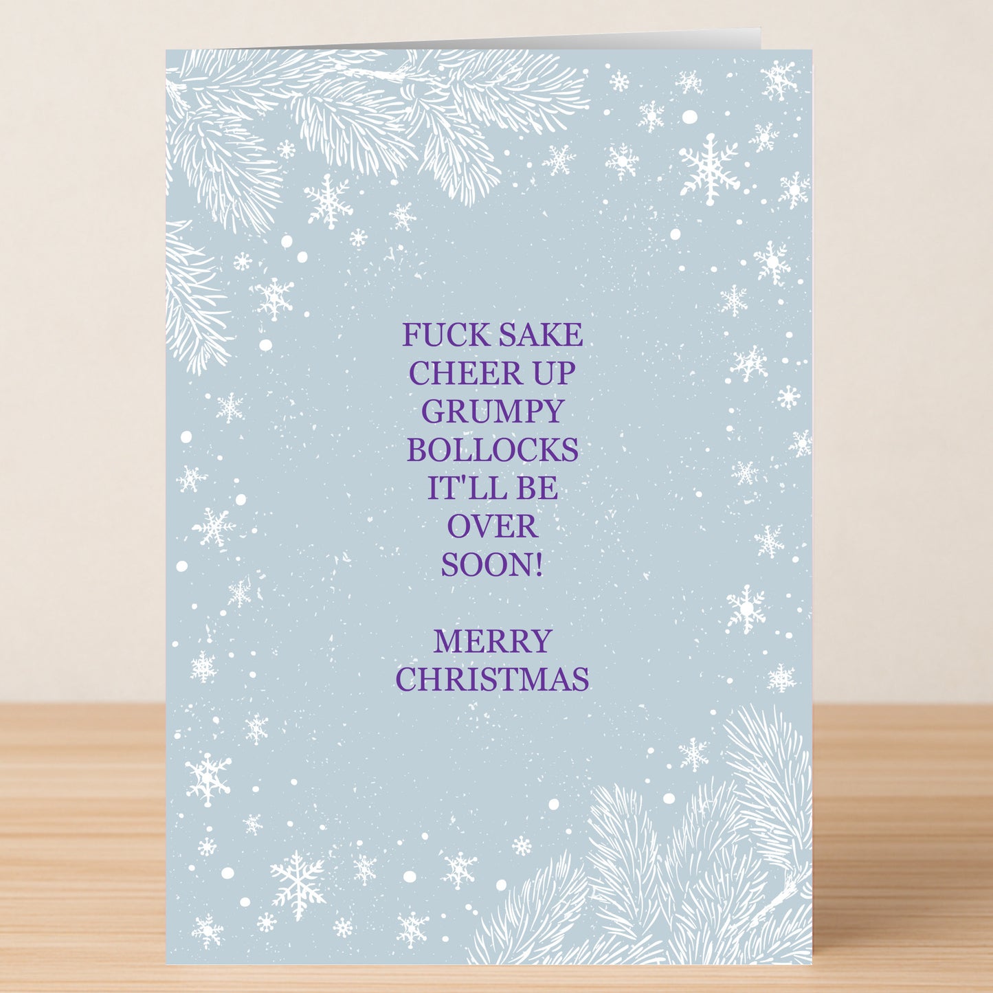 The Cheer Up Christmas Card by Twisted Gifts is a light blue card with white snowflakes and pine branches, featuring a humorous purple message that encourages someone to cheer up this holiday season.