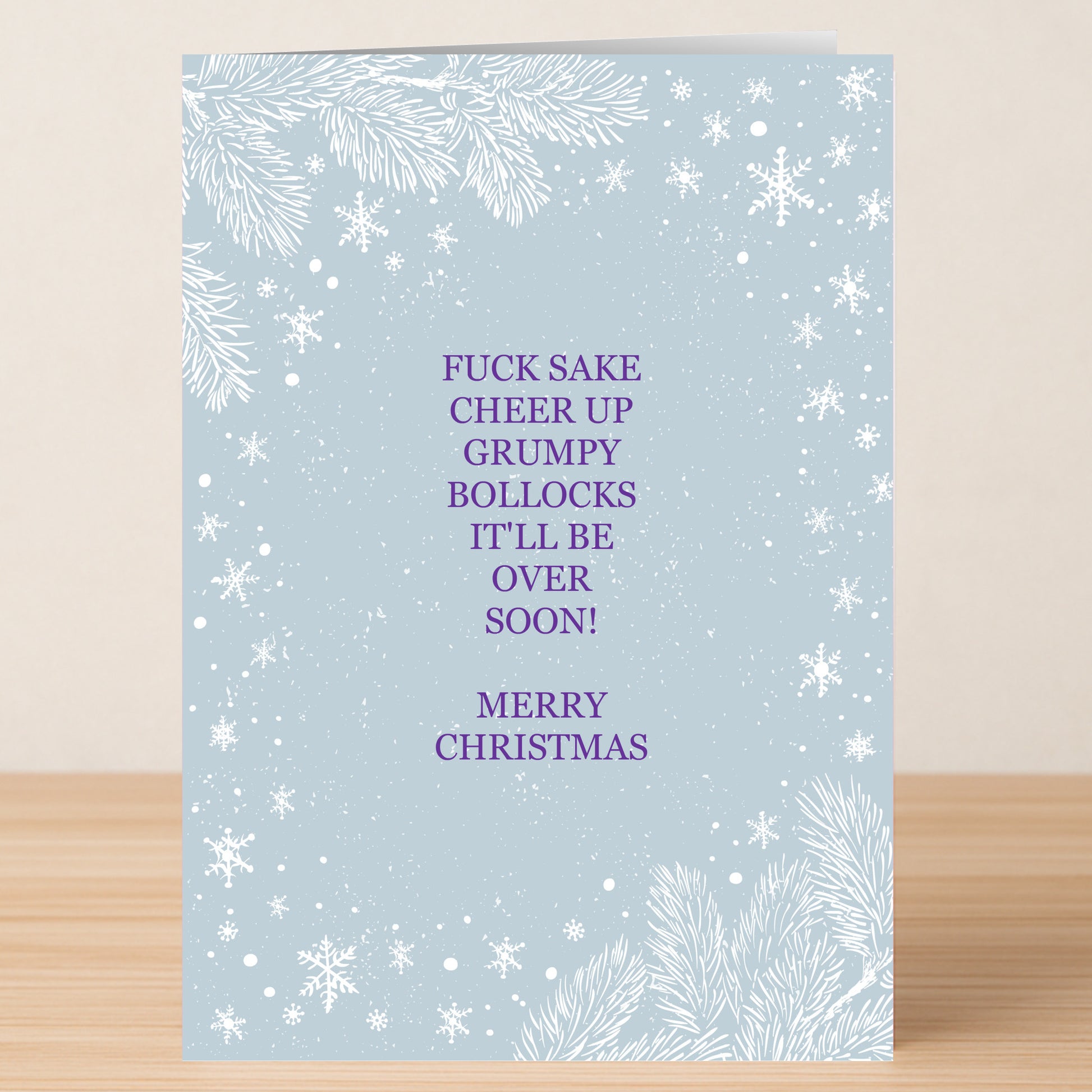 The Cheer Up Christmas Card by Twisted Gifts is a light blue card with white snowflakes and pine branches, featuring a humorous purple message that encourages someone to cheer up this holiday season.