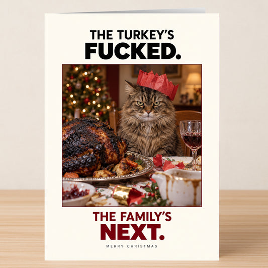 The Family Is Next - Funny Christmas Card features a grumpy long-haired cat in a red party hat sitting with a burnt turkey, captioned: “THE TURKEY’S FUCKED. THE FAMILY’S NEXT. MERRY CHRISTMAS.”