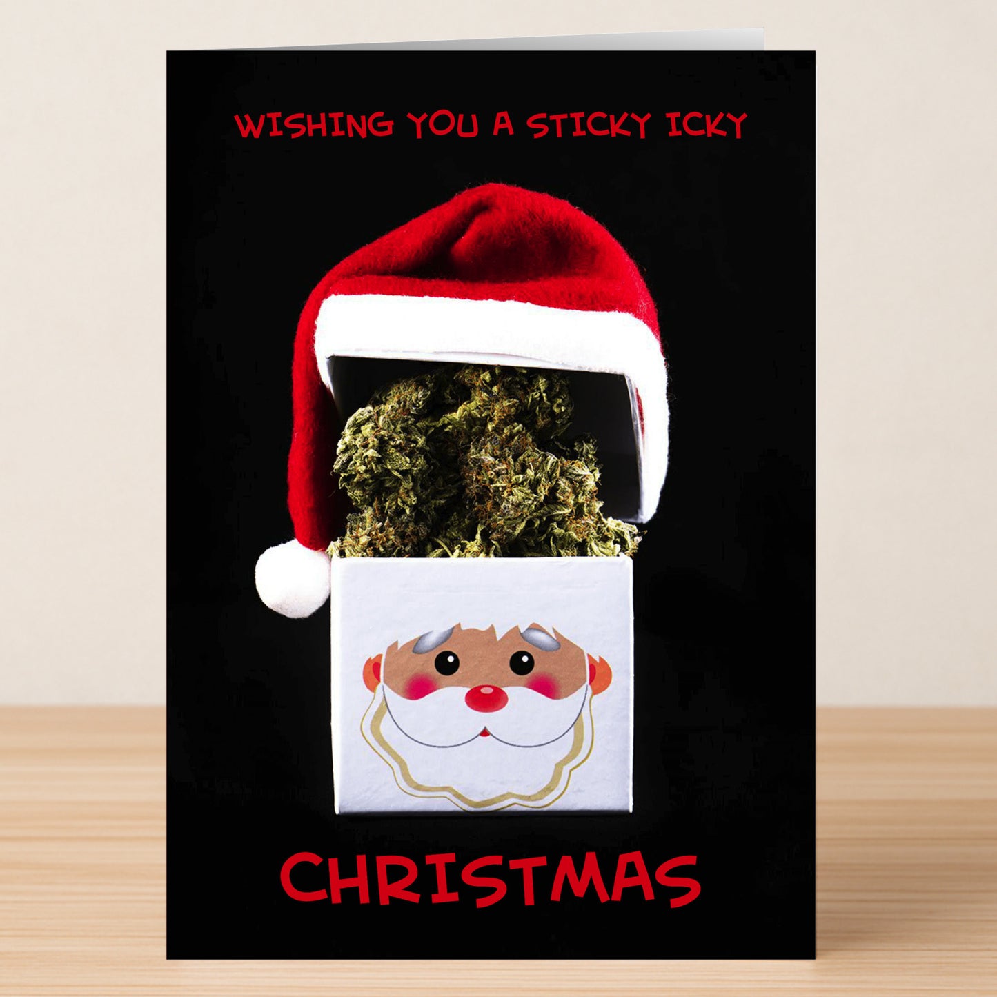 The Sticky Funny Christmas Card by Twisted Gifts features a box of cannabis with a Santa hat and cartoon face, plus the phrase "Wishing you a sticky icky Christmas." Send some holiday cheer with this humorous card!.