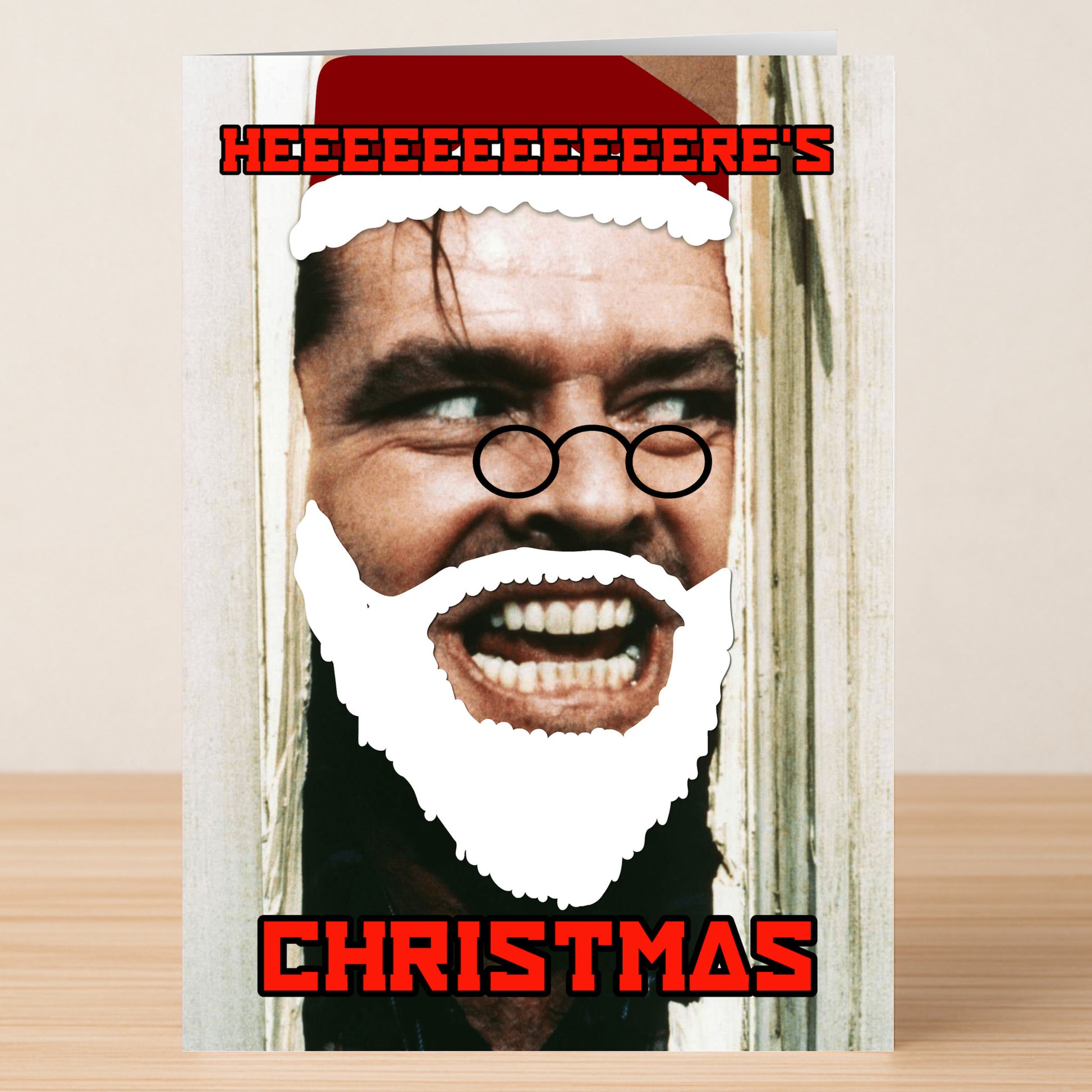 The Here's Christmas Funny Christmas Card by Twisted Gifts features a man's face at a broken door, edited with a Santa hat, glasses, and beard. The card reads "HEEEEEEEERE'S CHRISTMAS," making it perfect for the holiday season!.