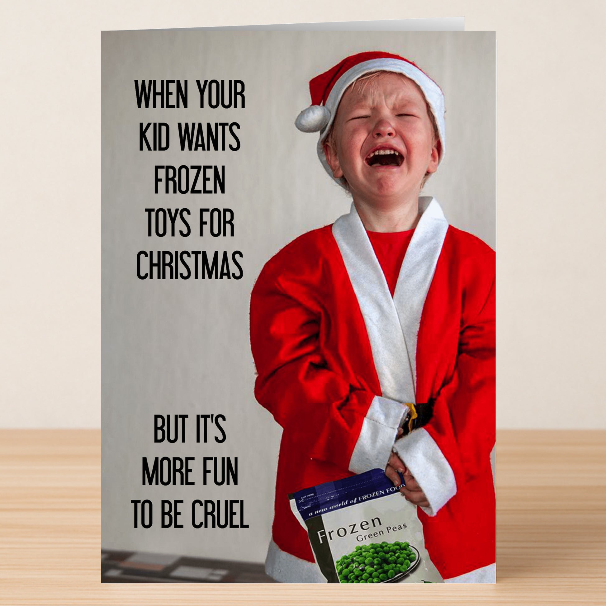 A child in a Santa costume cries while holding frozen peas. Text: "When your kid wants Frozen toys for Christmas, but it's more fun to be cruel." Frozen Funny Christmas Card by Twisted Gifts—perfect for your hilarious holiday greetings!.