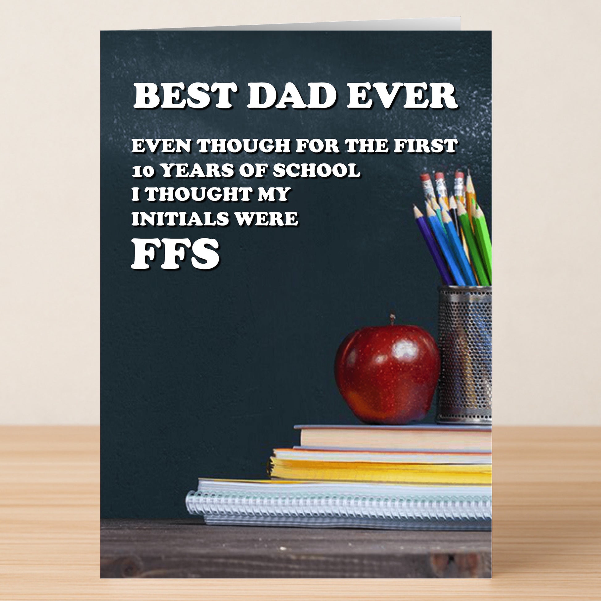 Twisted Gifts’ FFS Funny Father’s Day Card features quirky text—“Best Dad Ever...I thought my initials were FFS”—with stacked notebooks, an apple, and a pencil holder on a wooden surface. The perfect funny card for Dad!.