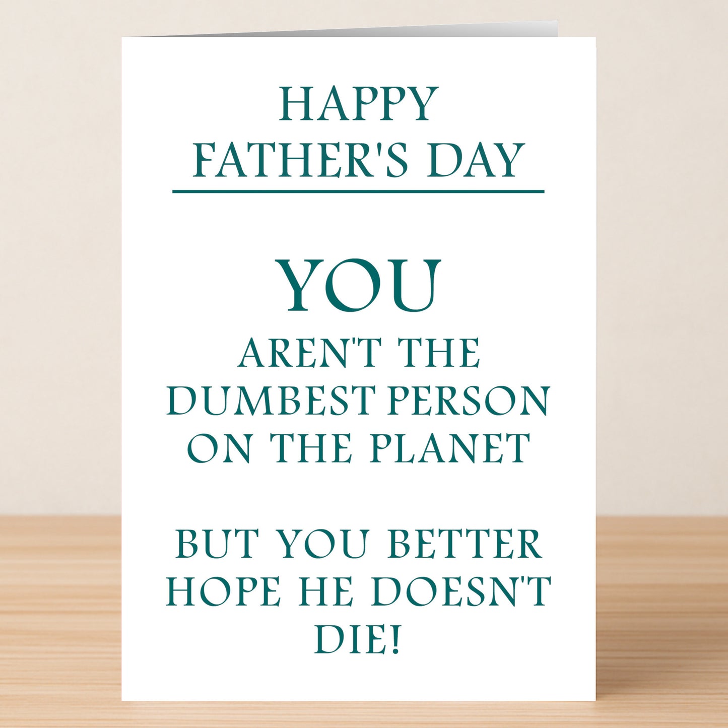 The Dumb Insulting Father's Day Card by Twisted Gifts sits on a wooden surface, featuring the playful message: "Happy Father's Day. You aren't the dumbest person on the planet, but you better hope he doesn't die!.