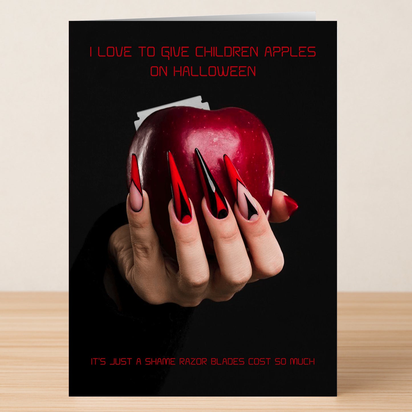 The Apples Funny Halloween Card by Twisted Gifts features a hand with long red and black nails holding an apple with razor blades—a humorous Halloween card poking fun at the classic "razor blade in apples" joke.