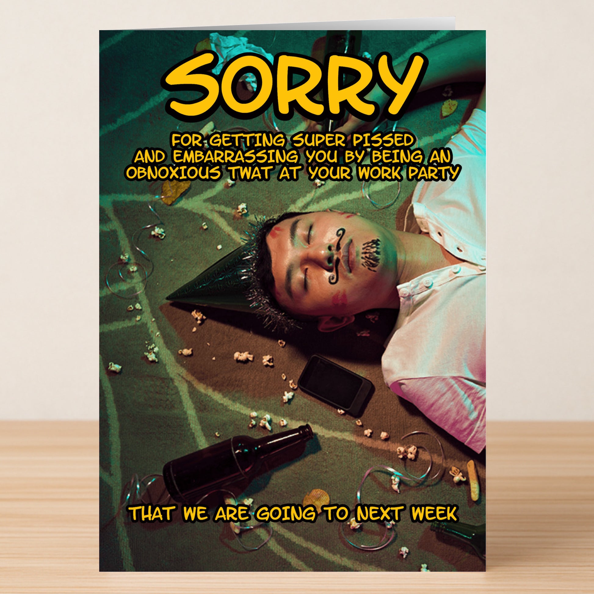 A man in a party hat lies on the floor with doodles on his face, beer bottles, popcorn, and a phone nearby. The Twisted Gifts Obnoxious Rude Sorry Card delivers a hilarious apology for being obnoxiously rude at the work party!.
