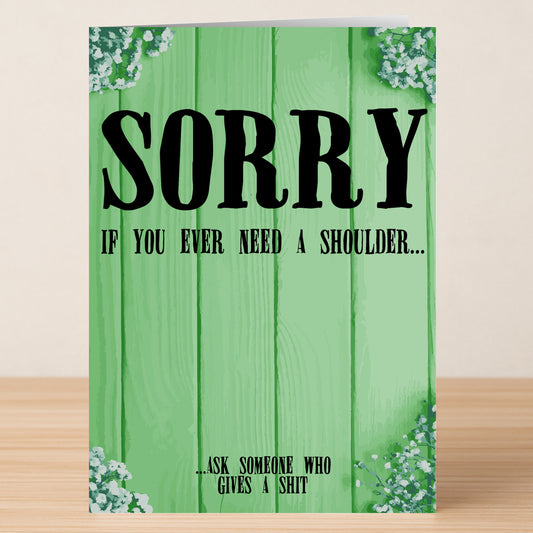 The Need A Shoulder Insulting Sorry Card by Twisted Gifts features a green wooden background, small white flowers in the corners, and reads: "SORRY if you ever need a shoulder... ...ask someone who gives a shit.