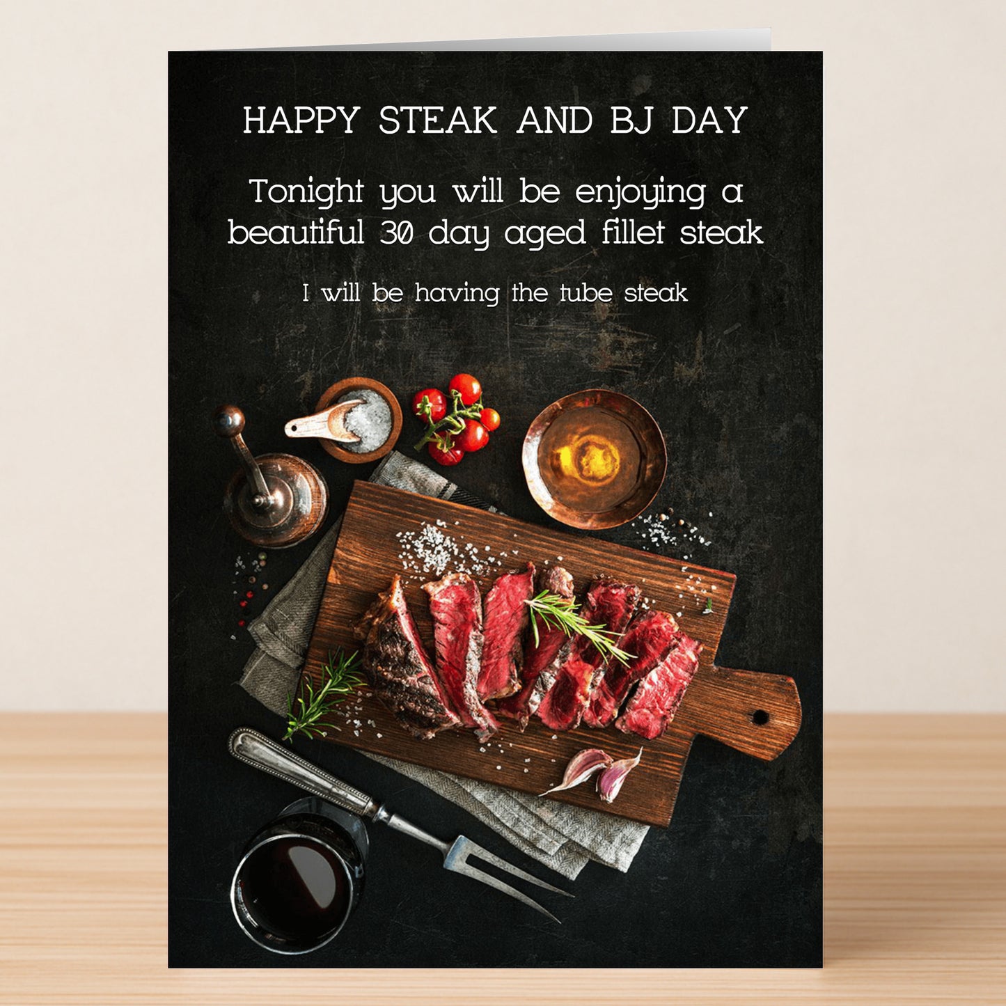 The Tube Steak Rude Steak And Blowjob Card by Twisted Gifts sits on a wooden table with sliced steak, condiments, and wine, displaying "Happy Steak and BJ Day" and a funny message.