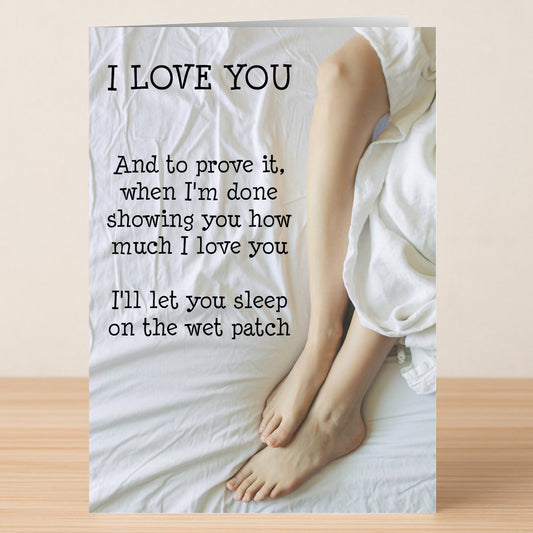 The Wet Patch Rude Valentine's Card by Twisted Gifts sits on a wooden surface, showing bare legs on white sheets with the cheeky message: "I LOVE YOU... I'll let you sleep on the wet patch.