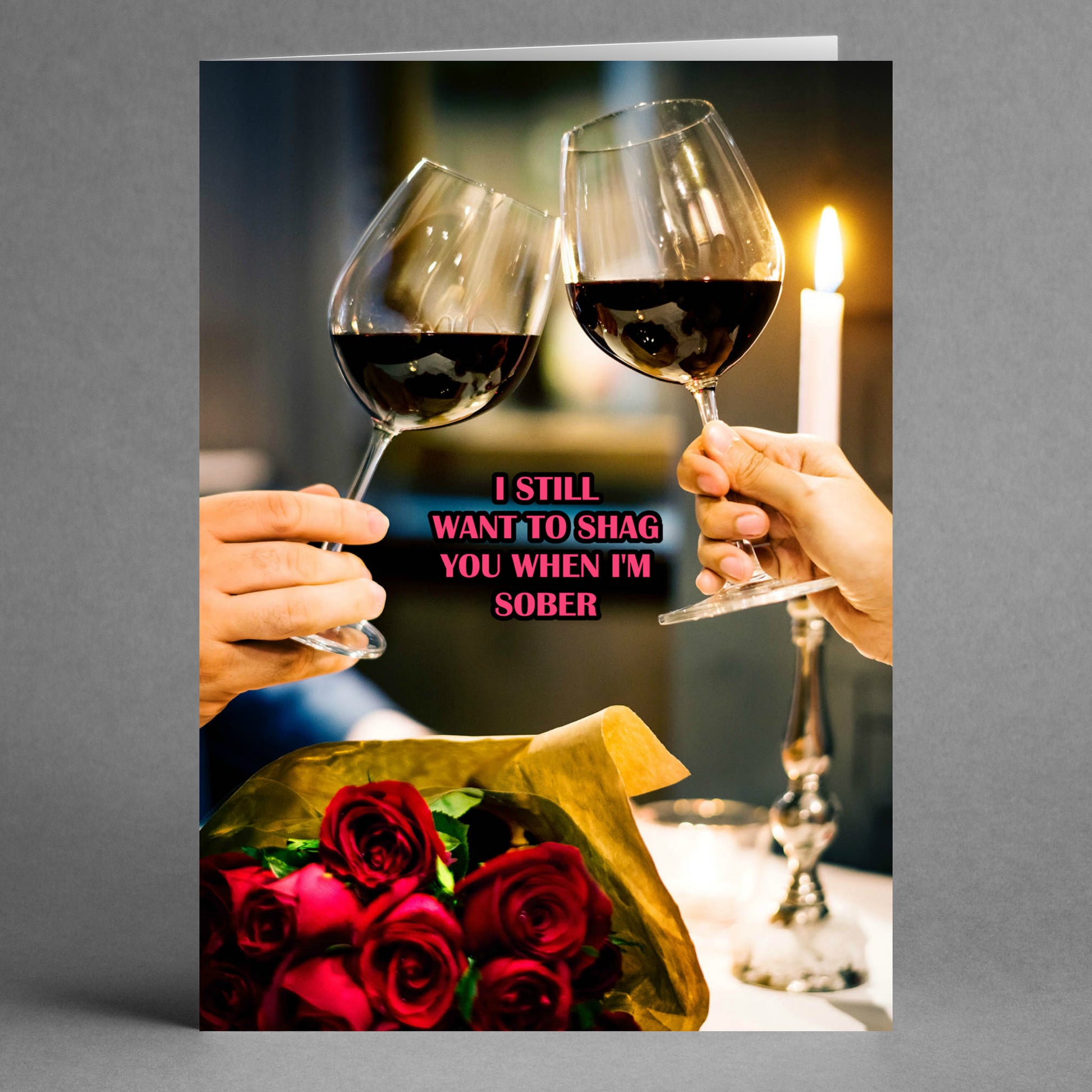 Two hands clink wine glasses over a bouquet of roses, with a candle glowing softly in the background. The text on the image says: "Sober Insulting Valentine's Card" by Twisted Gifts; a cheeky twist on a Valentine's Day greeting card that reads, "I still want to shag you when I'm sober.