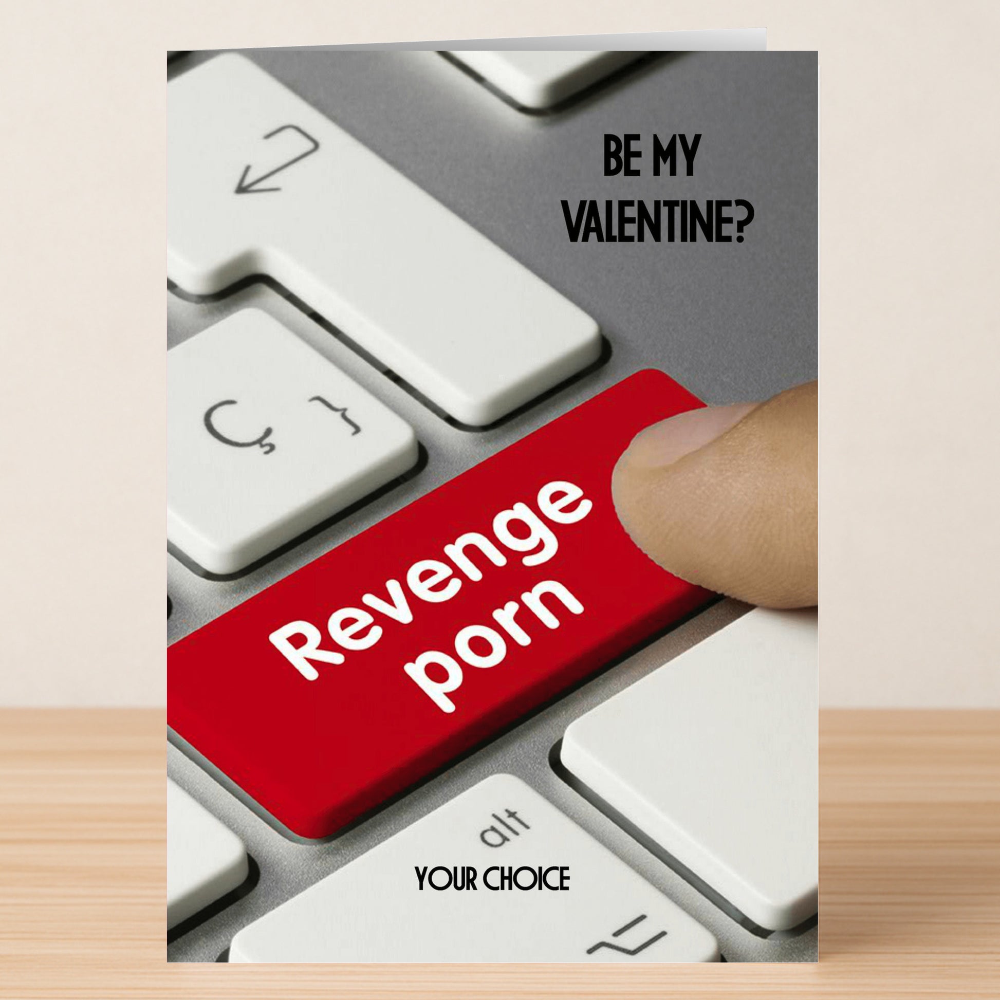 A finger presses a red keyboard key labeled "Revenge porn" beside "Be my Valentine?" and "Your choice," illustrating the Twisted Gifts Your Choice Insulting Valentine's Card—a card with a truly twisted sense of humor.