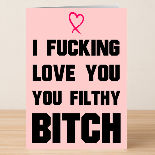 The Filthy Bitch Rude Greeting Card by Twisted Gifts is a pink card with a heart and bold black text saying, "I fucking love you you Filthy Bitch," shown on a wooden surface.