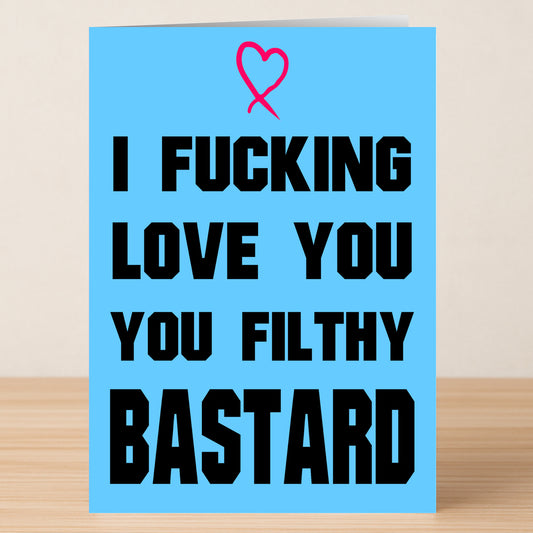 The Filthy Bastard Rude Greeting Card by Twisted Gifts features a blue design with a pink heart and bold black text, "I FUCKING LOVE YOU YOU FILTHY BASTARD." This Valentine’s card stands on a wooden surface.