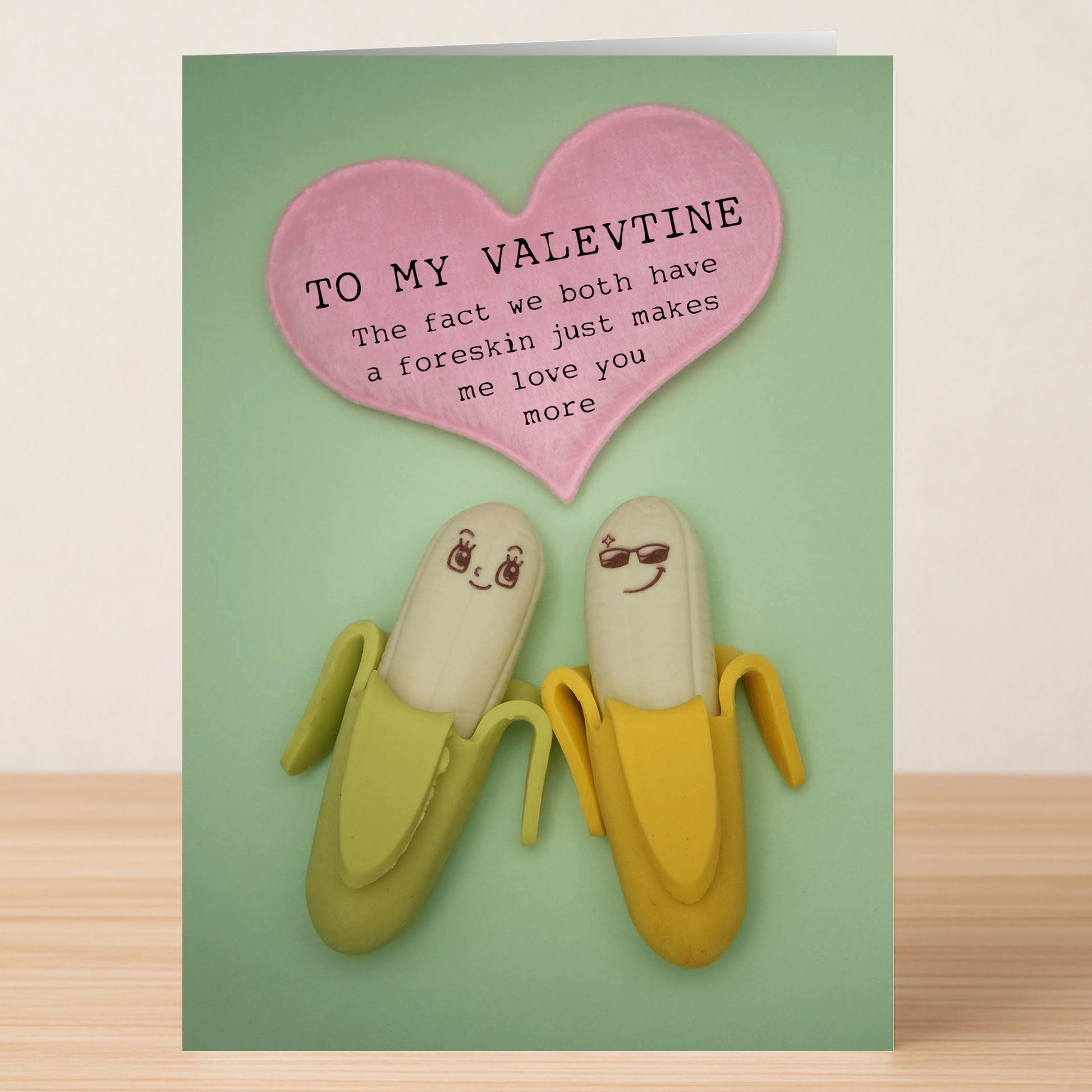 Two bananas with cute faces sit under a pink heart reading, "TO MY VALENTINE: The fact we both have a foreskin just makes me love you more." A cheeky twist from Twisted Gifts' Both Of Us Insulting Valentine's Card.