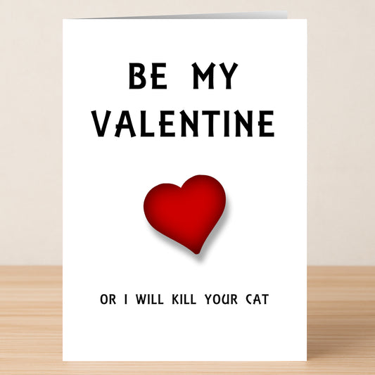 The Cat Gets It Twisted Valentine's Card by Twisted Gifts features "BE MY VALENTINE" above a red heart and the cheeky message "OR I WILL KILL YOUR CAT," displayed on a wooden surface.