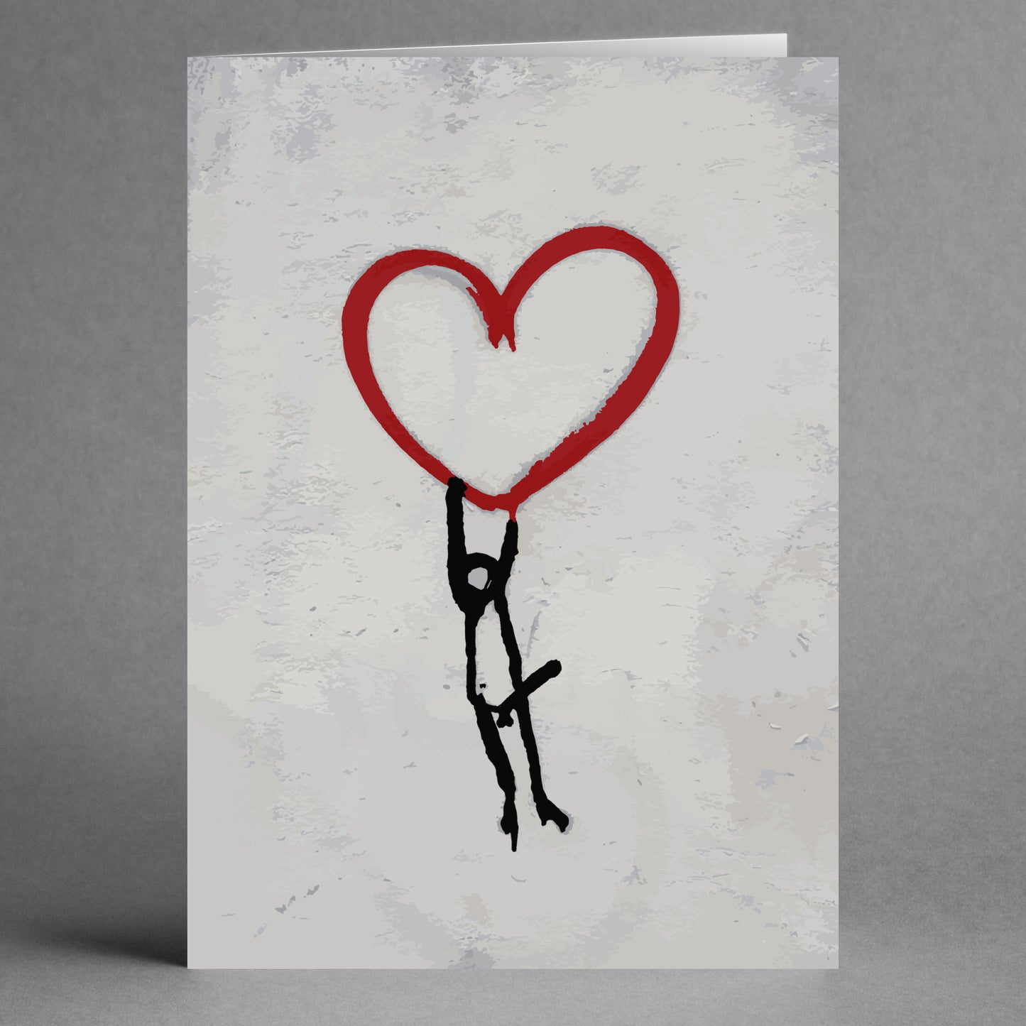 The Banksy Style Funny Valentine's Card by Twisted Gifts features a comical stick figure dangling from a big red heart, set against a textured gray backdrop, making it ideal for a humorous Valentine's gesture.