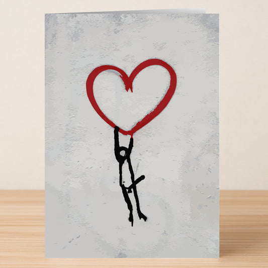 The Banksy Style Funny Valentine's Card by Twisted Gifts features a stick figure humorously hanging from a big red heart on a light gray background, displayed on a wooden surface.