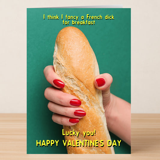 A hand with red nail polish holds a baguette against a green background on Twisted Gifts' French Dick Rude Valentine's Card, which reads: “I think I fancy a French dick for breakfast. Lucky you! Happy Valentine’s Day.”.