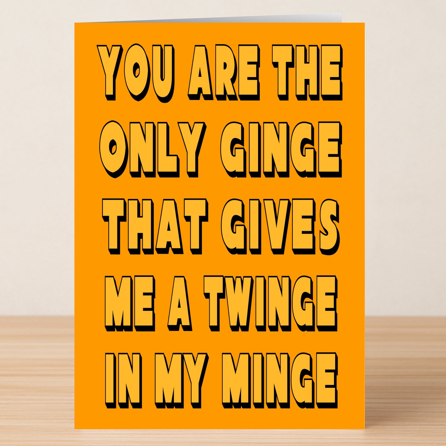 The Twinge Funny Valentine's Card by Twisted Gifts features bold black text on orange: "You are the only ginge that gives me a twinge in my minge." A perfect limited edition card for someone special, shown on a wooden surface.