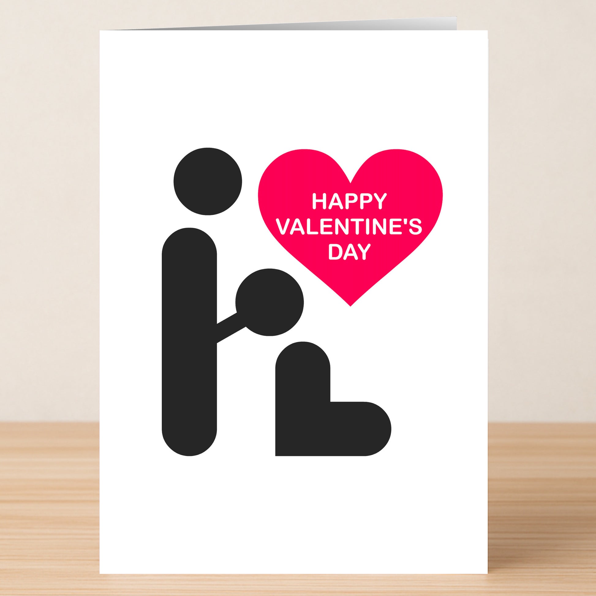 The Simple BJ Rude Valentine's Card by Twisted Gifts features minimalist figures—one kneeling before another with a pink heart above that says "HAPPY VALENTINE'S DAY." Its cheeky, humorous design pops on a wooden surface.