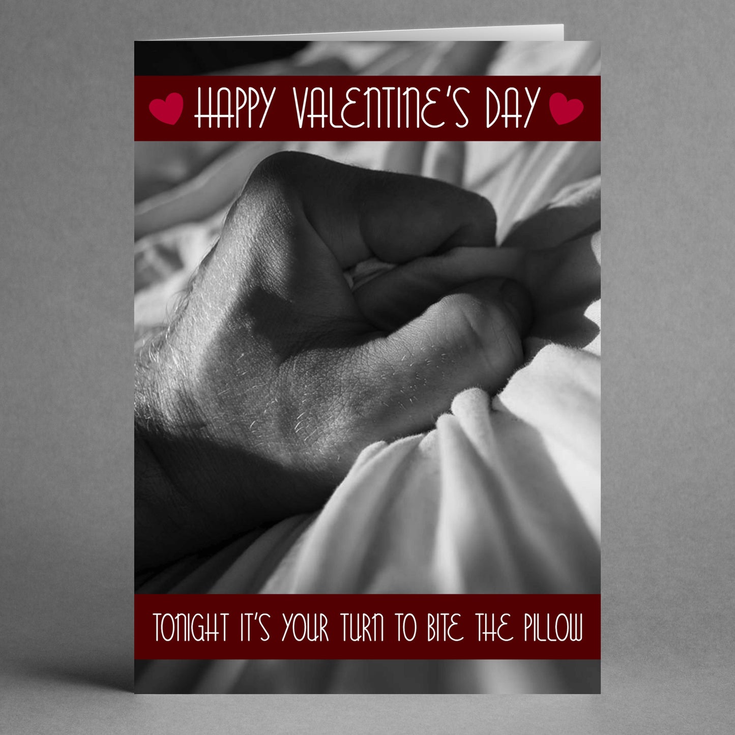 A black and white image of a hand clutching a pillow is accompanied by the playful message, "Happy Valentine's Day" and the cheeky line, "Tonight it's your turn to bite the pillow." The card is bordered in red with heart symbols, adding a touch of flair to this daring Pillow Rude Valentine's Card by Twisted Gifts.