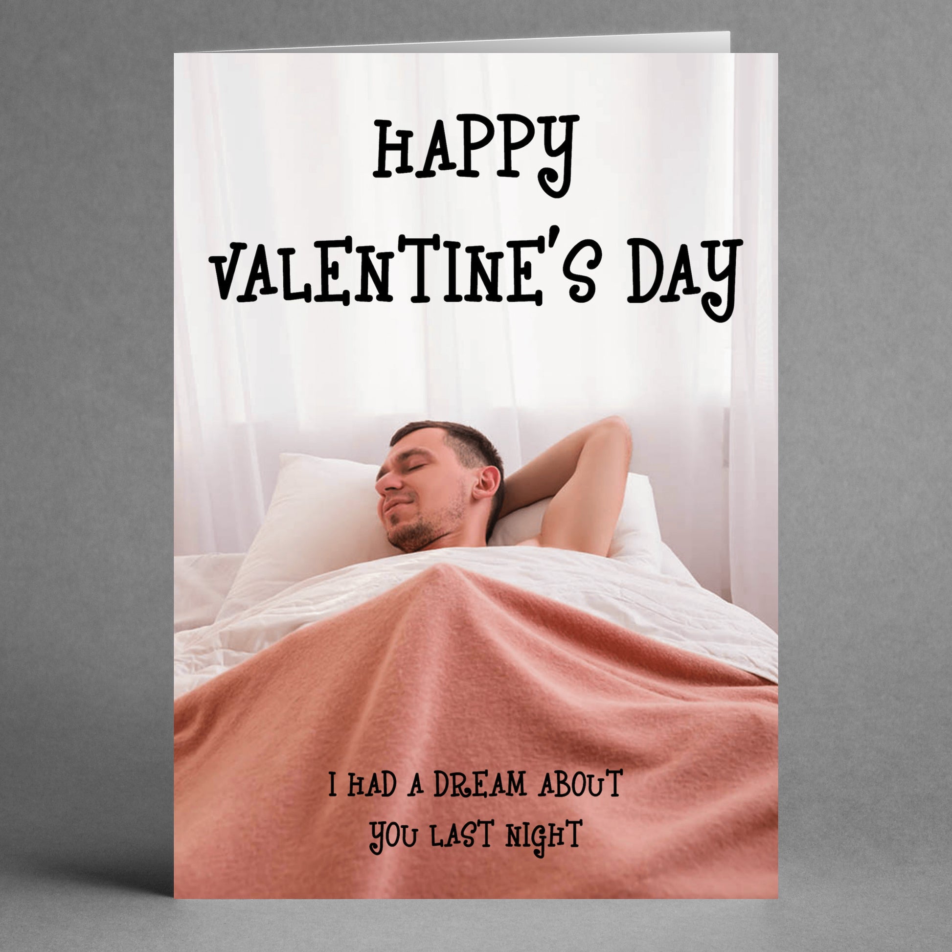 A man sleeps in bed wrapped in a pink blanket. Beside him, a Twisted Gifts Dream About You Rude Valentine's Card quietly announces, "Happy Valentine's Day," and cheekily adds, "I had a dream about you last night," infusing sweet dreams into the holiday.