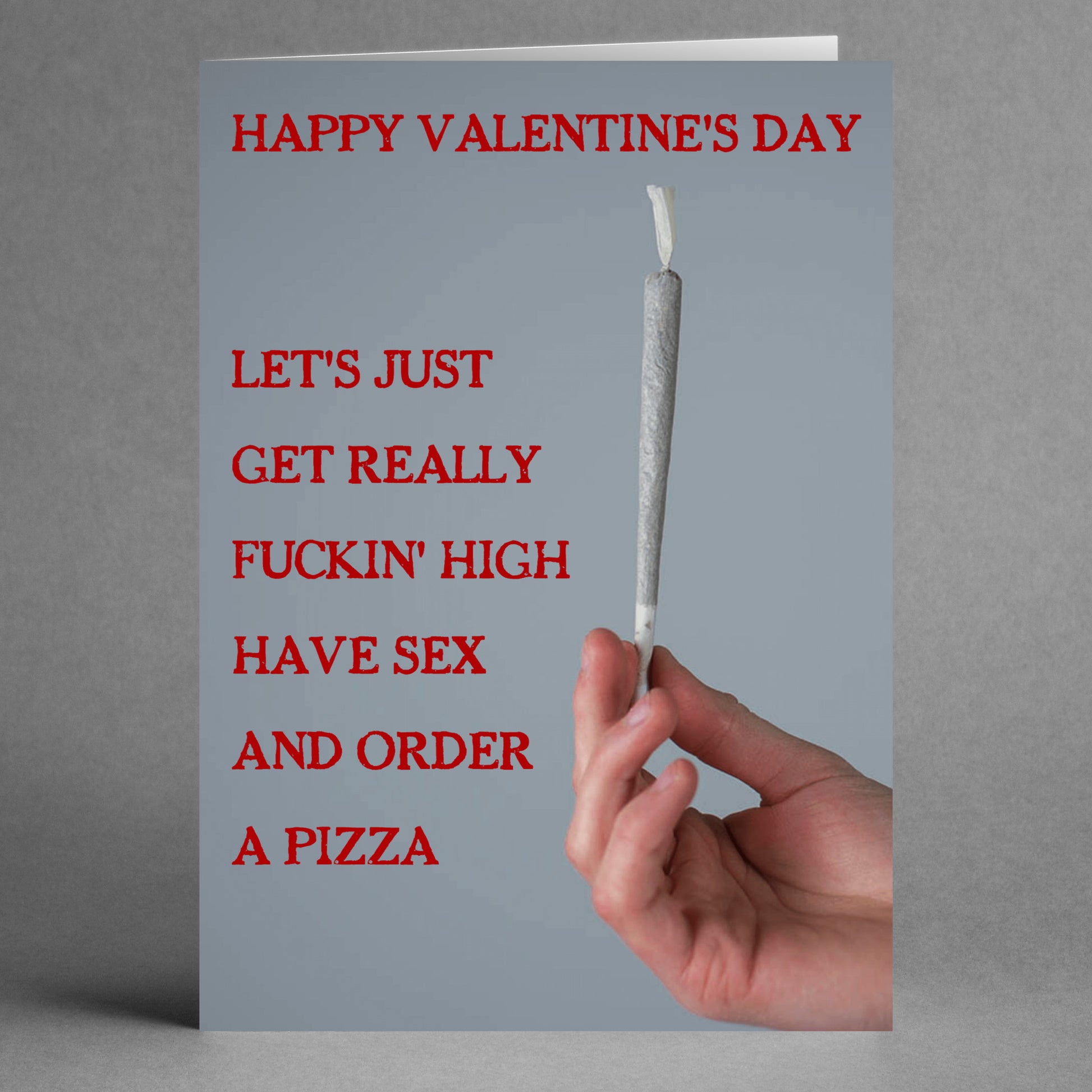 A playful card from Twisted Gifts, the Pizza Rude Valentine's Card, features an illustration of a hand holding a rolled joint against a gray background. The bold text states: "Happy Valentine's Day. Let's just get really fuckin' high, have sex, and order a pizza." It's ideal for those who enjoy an unabashedly fun and daring sense of humor!