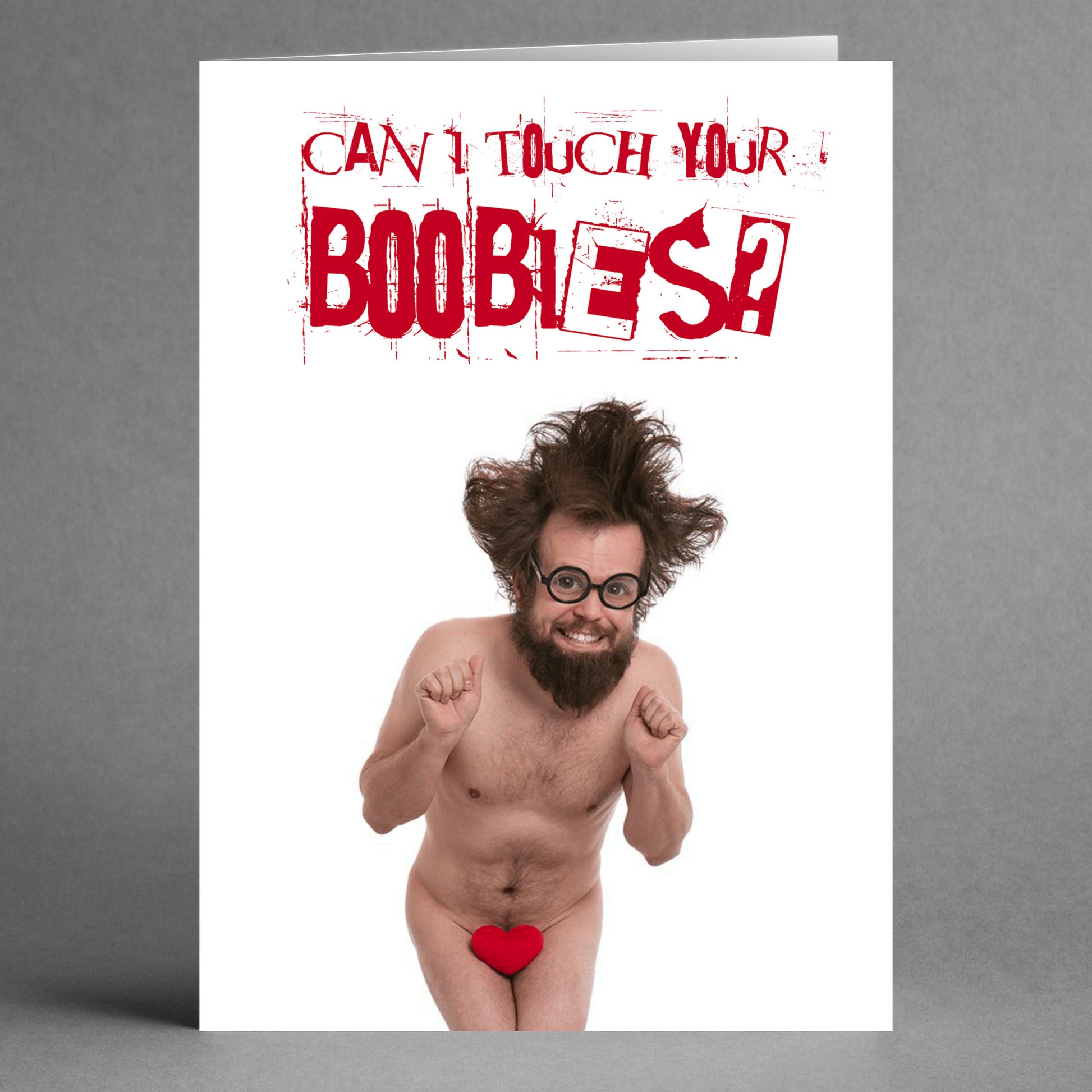 A man with glasses and messy hair is jumping, holding a small red heart for cover. Above him, the text says, "Can I touch your boobies?" This humorous Valentine's Day card from Twisted Gifts is aptly named the Boobies Rude Greeting Card.