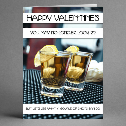 A card from Twisted Gifts reads: "Happy Valentine's. You may no longer look 22, but let's see what a couple of shots can do." It's ideal for those with a sense of humor looking to make this a memorable Valentine's Day. Below the card, two shot glasses with lemon wedges rest on the table.