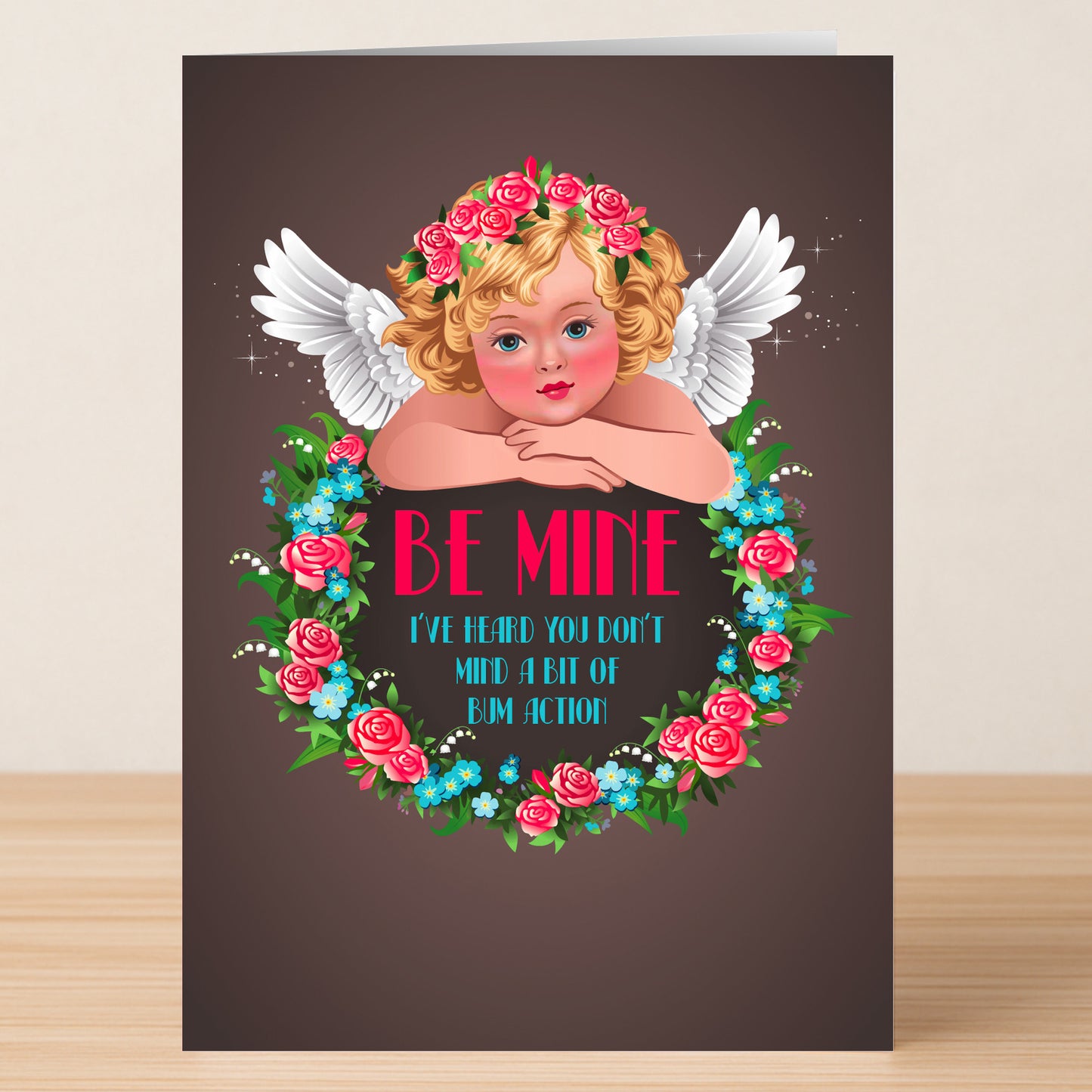 The Action Rude Valentine's Card by Twisted Gifts features a cheeky cherub with wings, a floral crown, a rose wreath, and the playful message: "Be Mine. I've heard you don't mind a bit of bum action.