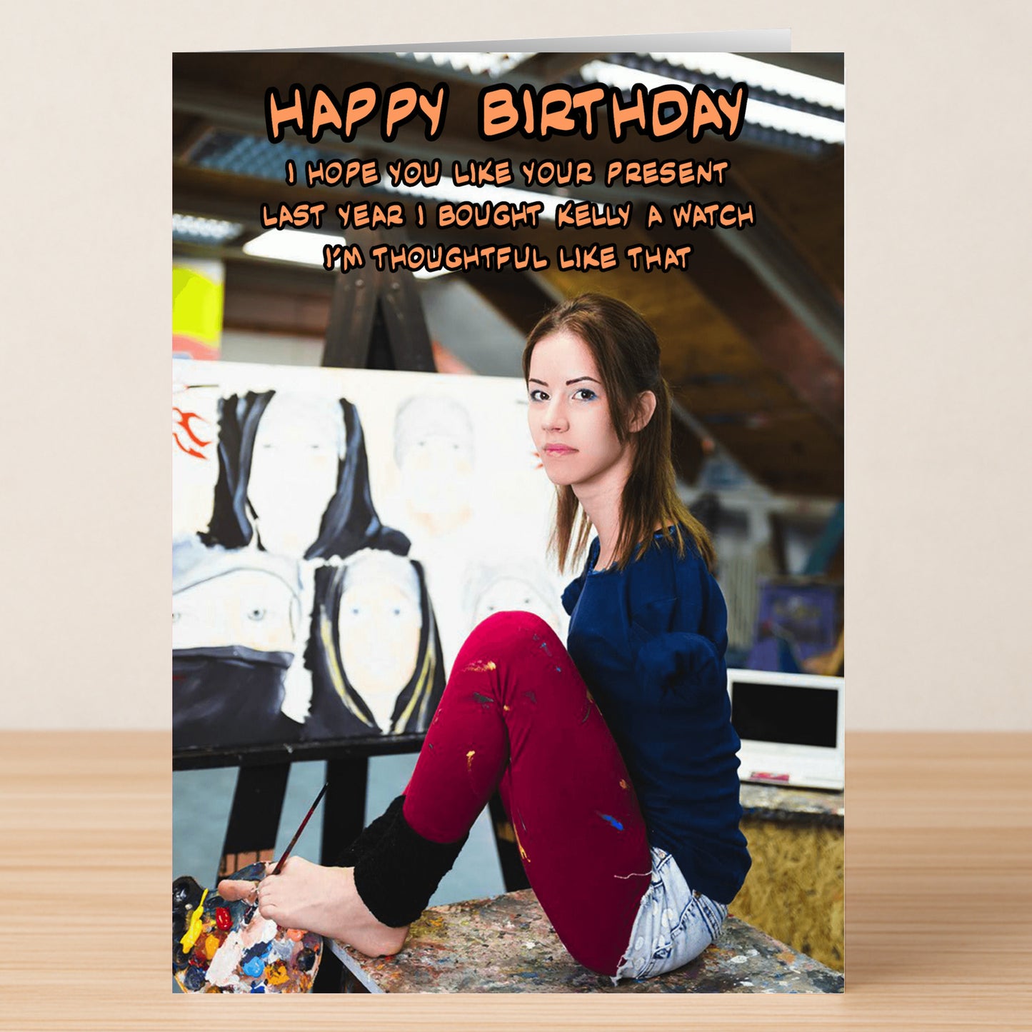 A woman with prosthetic legs sits on a stool by an easel of paintings; above her is the “Thoughtful Gift” joke from Twisted Gifts’ Funny Rude Birthday Card—a dark humour birthday message about buying Kelly a watch.