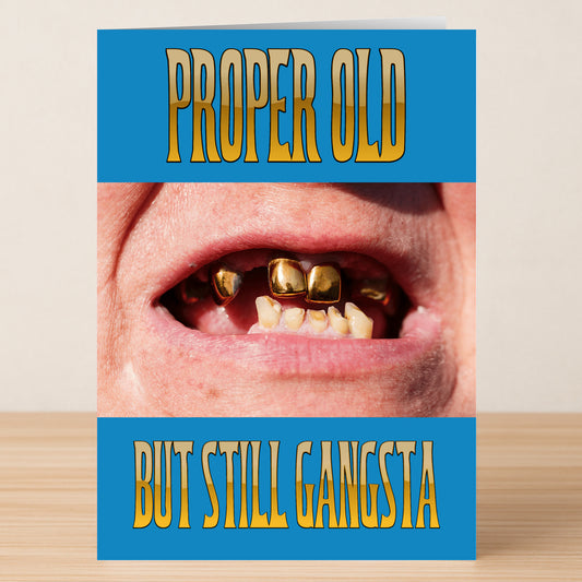 The Still Gangsta Funny Birthday Card by Twisted Gifts features a close-up of an older person's mouth with gold teeth, framed by blue panels with yellow "PROPER OLD" and "BUT STILL GANGSTA" text—a perfect choice for anyone who loves humor!.