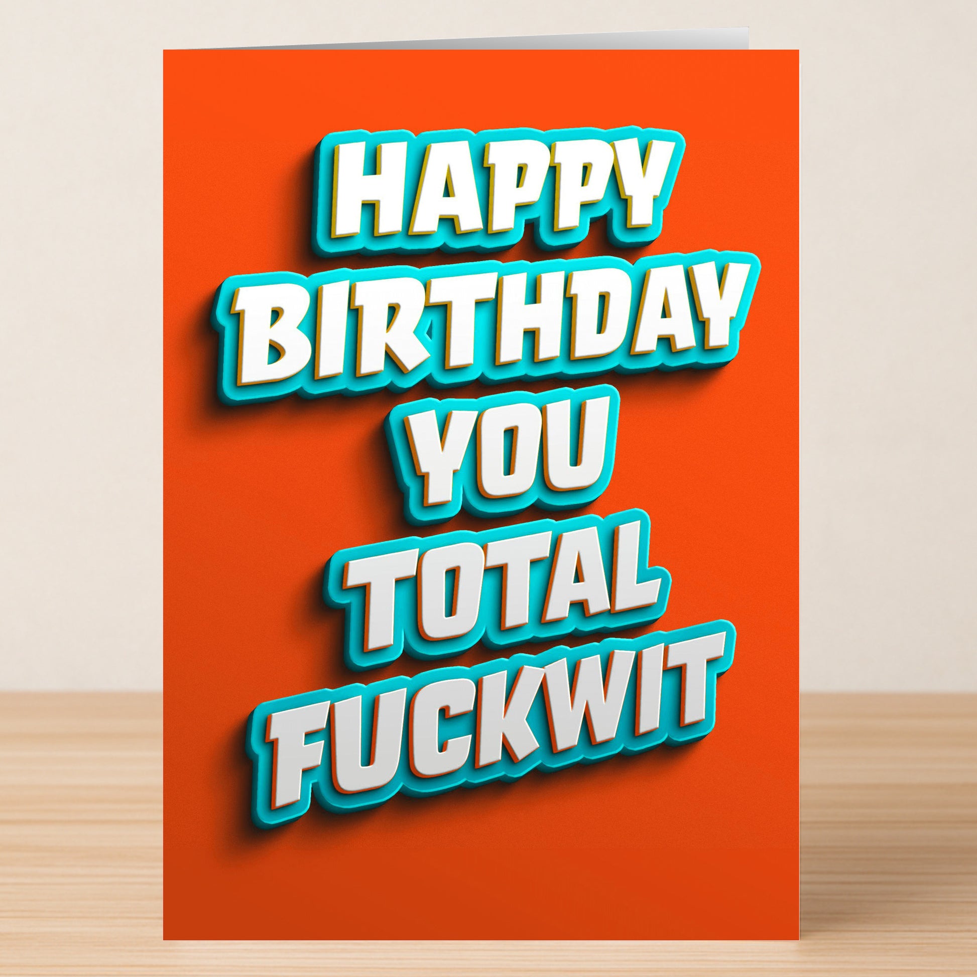 Twisted Gifts’ “Total Fuckwit – Insulting Birthday Card” features bold white and blue text on an orange background reading, "Happy Birthday You Total Fuckwit," and is shown standing on a wooden surface.
