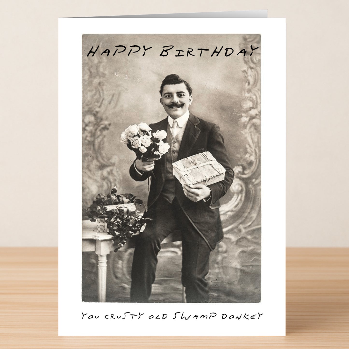 The Swamp Donkey – Insulting Birthday Card by Twisted Gifts features a black-and-white photo of a man in a suit with flowers and a gift, plus the text "Happy Birthday" and "You crusty old swamp donkey." Perfect rude card for those who love edgy humor.