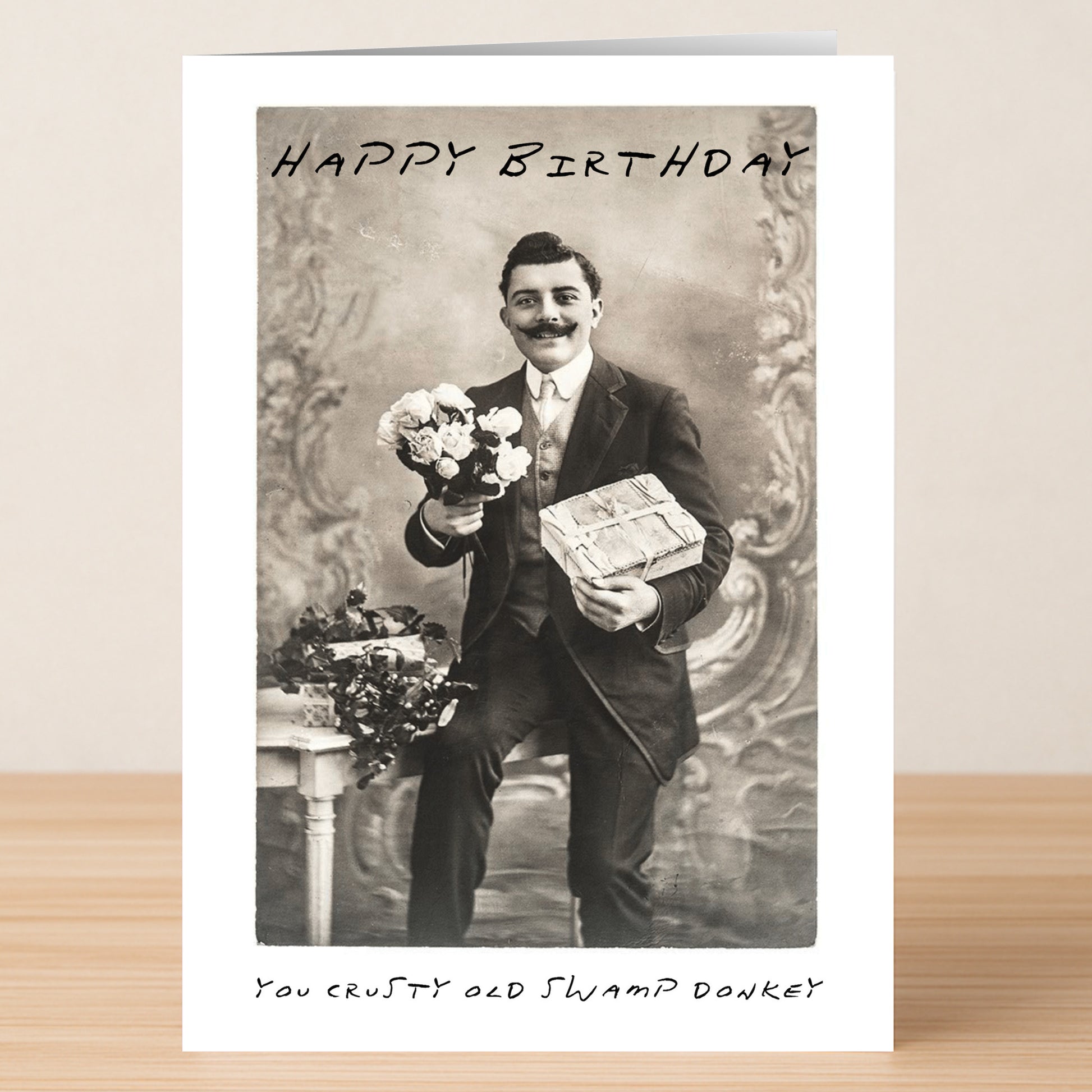 The Swamp Donkey – Insulting Birthday Card by Twisted Gifts features a black-and-white photo of a man in a suit with flowers and a gift, plus the text "Happy Birthday" and "You crusty old swamp donkey." Perfect rude card for those who love edgy humor.