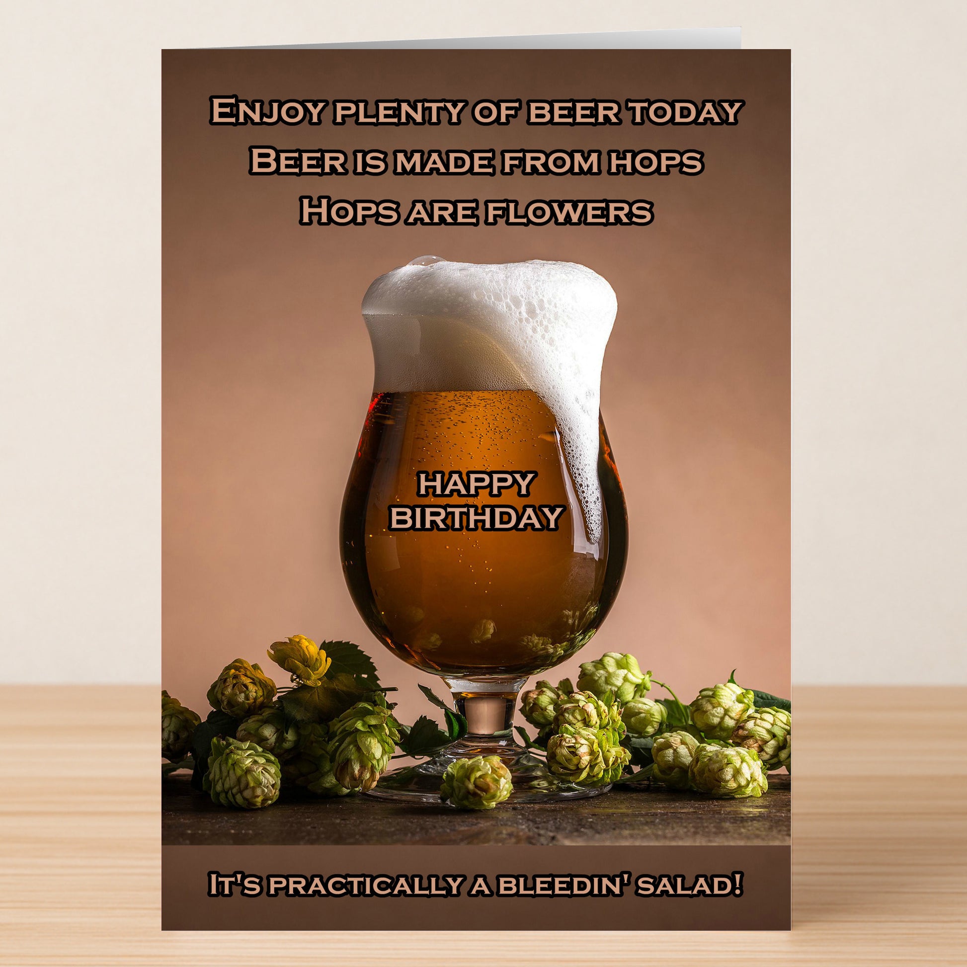 A glass of foamy beer sits among fresh hops beside Twisted Gifts' Funny Rude Birthday Card – “Salad” Beer Joke Card, featuring cheeky food humour about beer and hops counting as a salad.