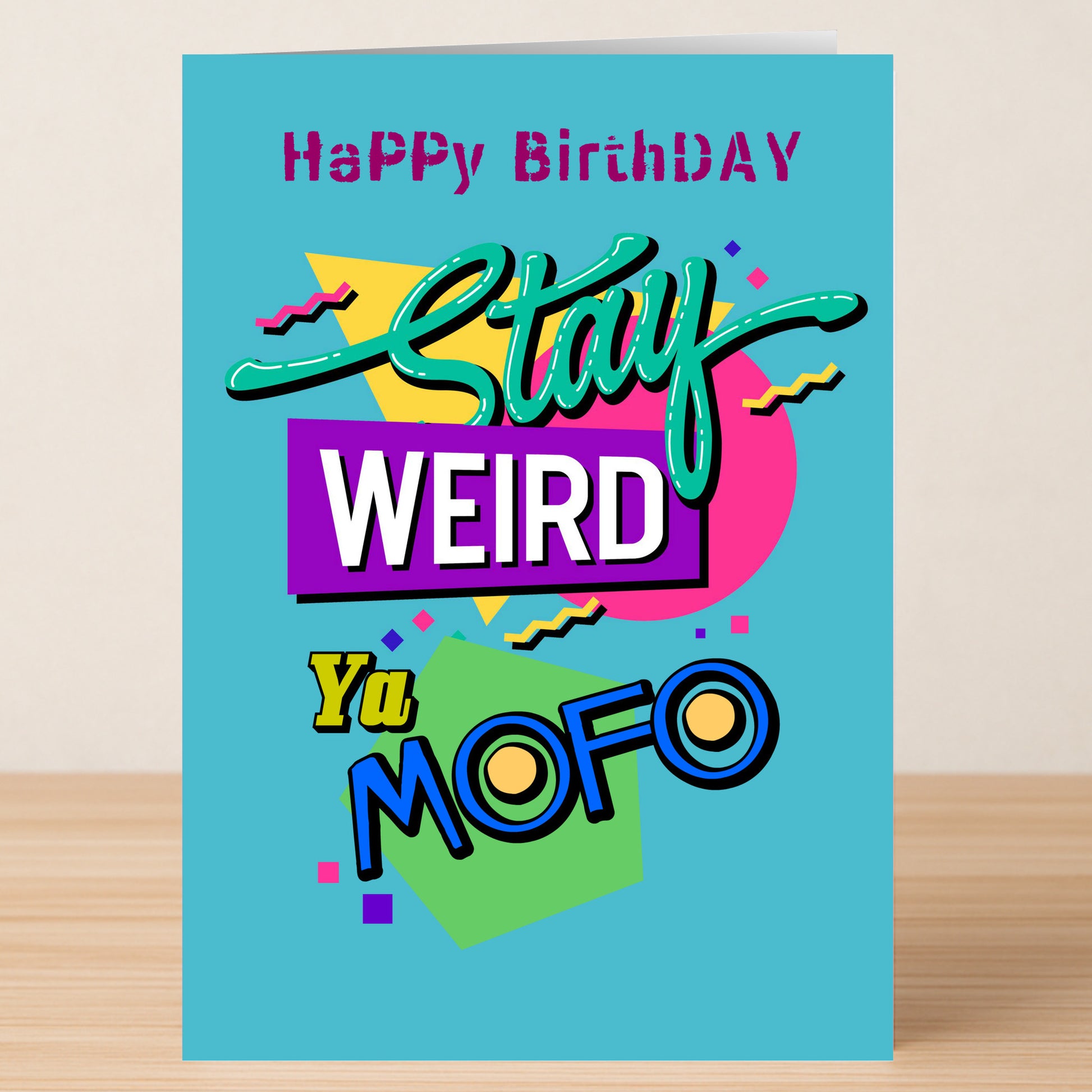 The Stay Weird – Funny Birthday Card by Twisted Gifts features colorful “Happy Birthday Stay Weird Ya MOFO” text on blue cardstock, shown on a wooden surface.