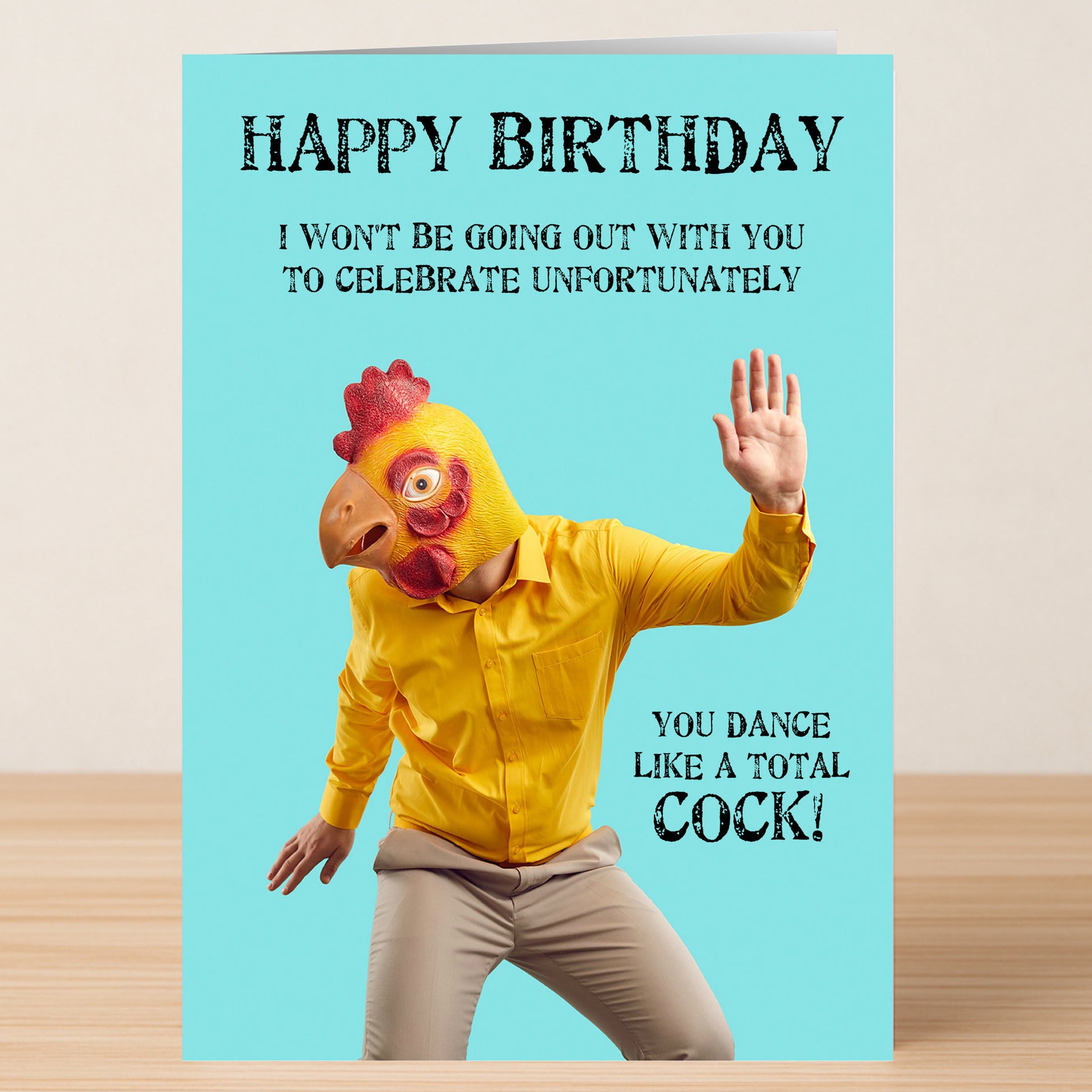 Wearing a chicken mask and yellow shirt, someone poses with Twisted Gifts’ "Total Cock – Insulting Birthday Card," which reads: "Happy Birthday. You dance like a total cock!.