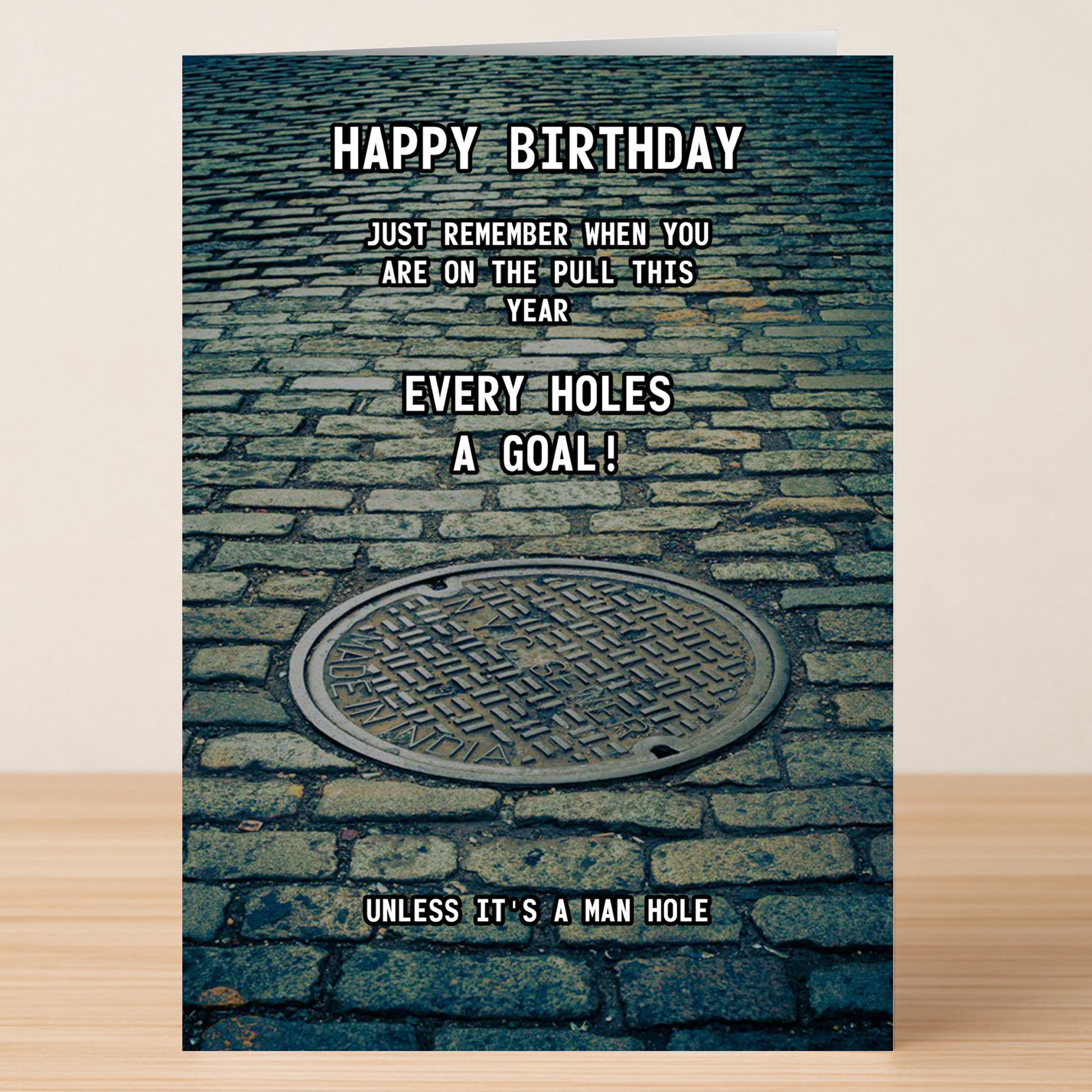 The Man Hole – Rude Birthday Card by Twisted Gifts features a manhole cover on cobblestone and the cheeky message: "Happy Birthday. Just remember when you are on the pull this year every hole's a goal! Unless it's a man hole.