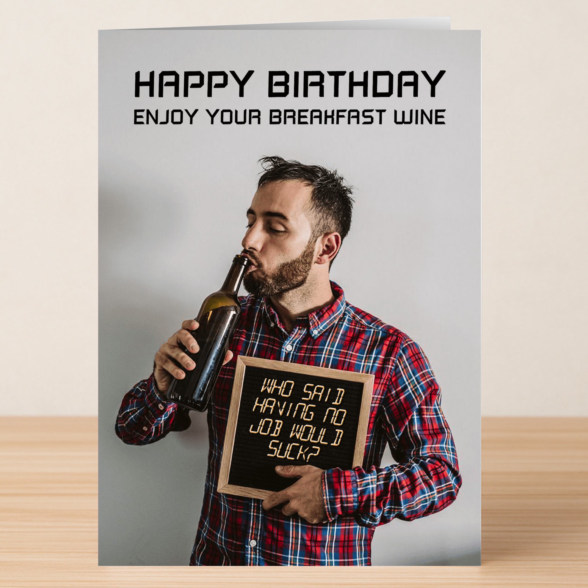 A man in a plaid shirt drinks wine and holds a sign saying, "Who said having no job would suck?" The Suck – Insulting Birthday Card by Twisted Gifts reads, "Happy Birthday. Enjoy your breakfast wine.
